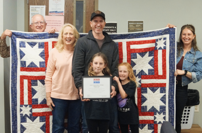 white man and woman holding up a red, white and blue quilt behind a white man with an older woman (mother) and his daughers